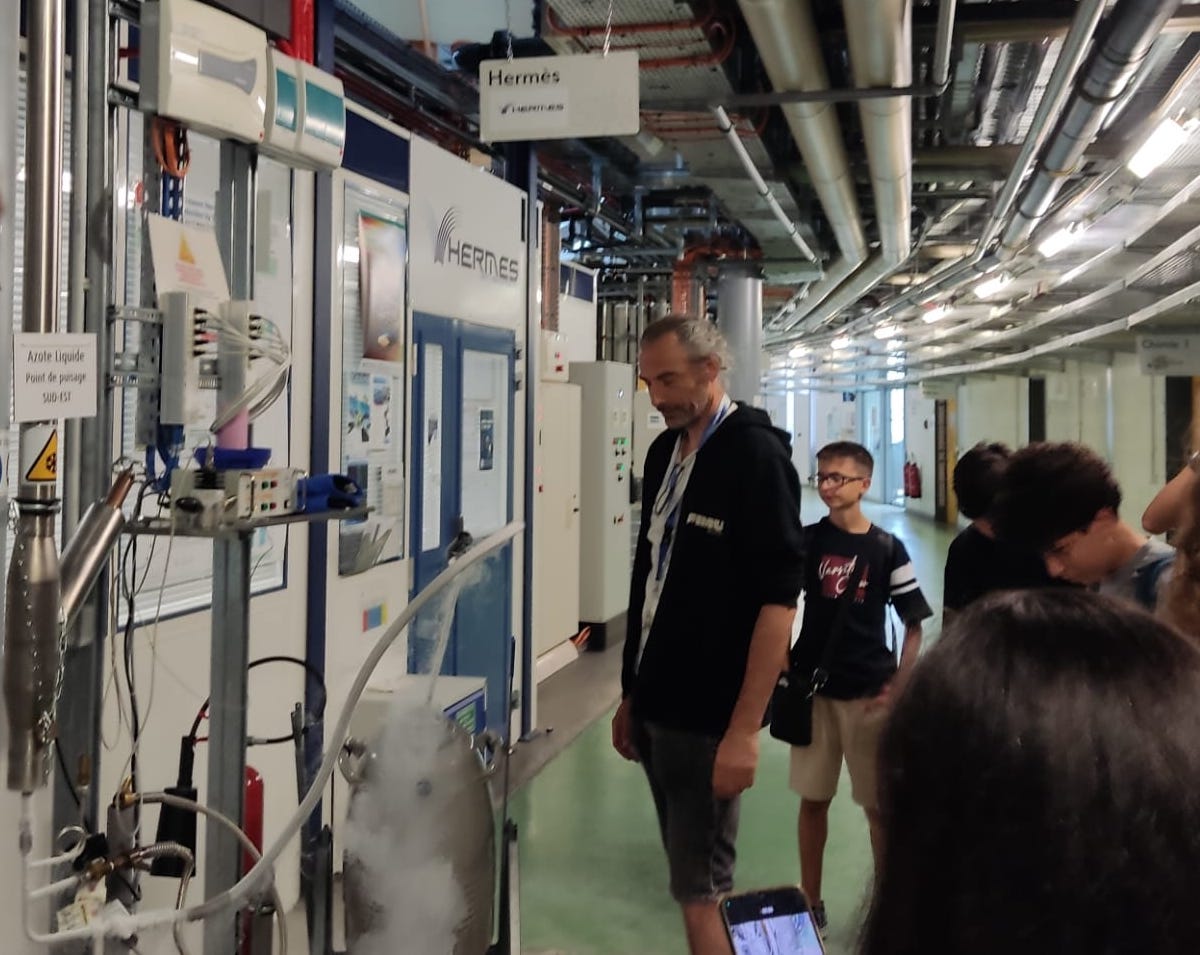 Un gruppo di studenti del progetto RIALE-EU visita una delle beam line del Sincrotrone SOLEIL a Parigi, guidati da un ricercatore. In primo piano, tubazioni e vapori di azoto liquido; sullo sfondo, un ambiente tecnologico con strutture e cartelli in francese.
